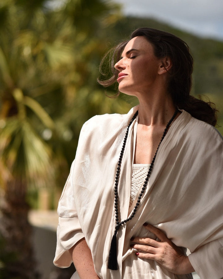 Woman in a white outfit with a long necklace standing outdoors with palm trees in the background
