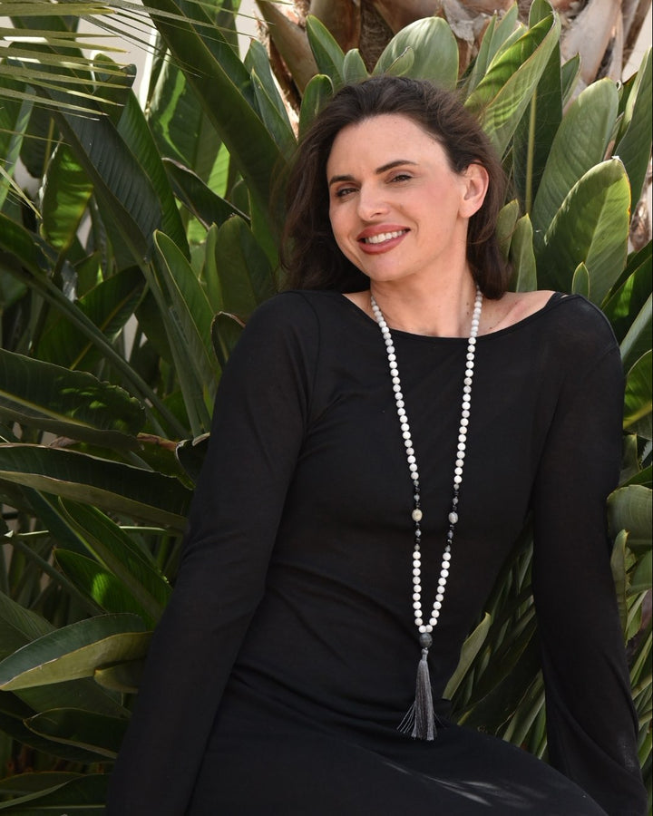 Woman in a black outfit with a long necklace sitting among green plants
