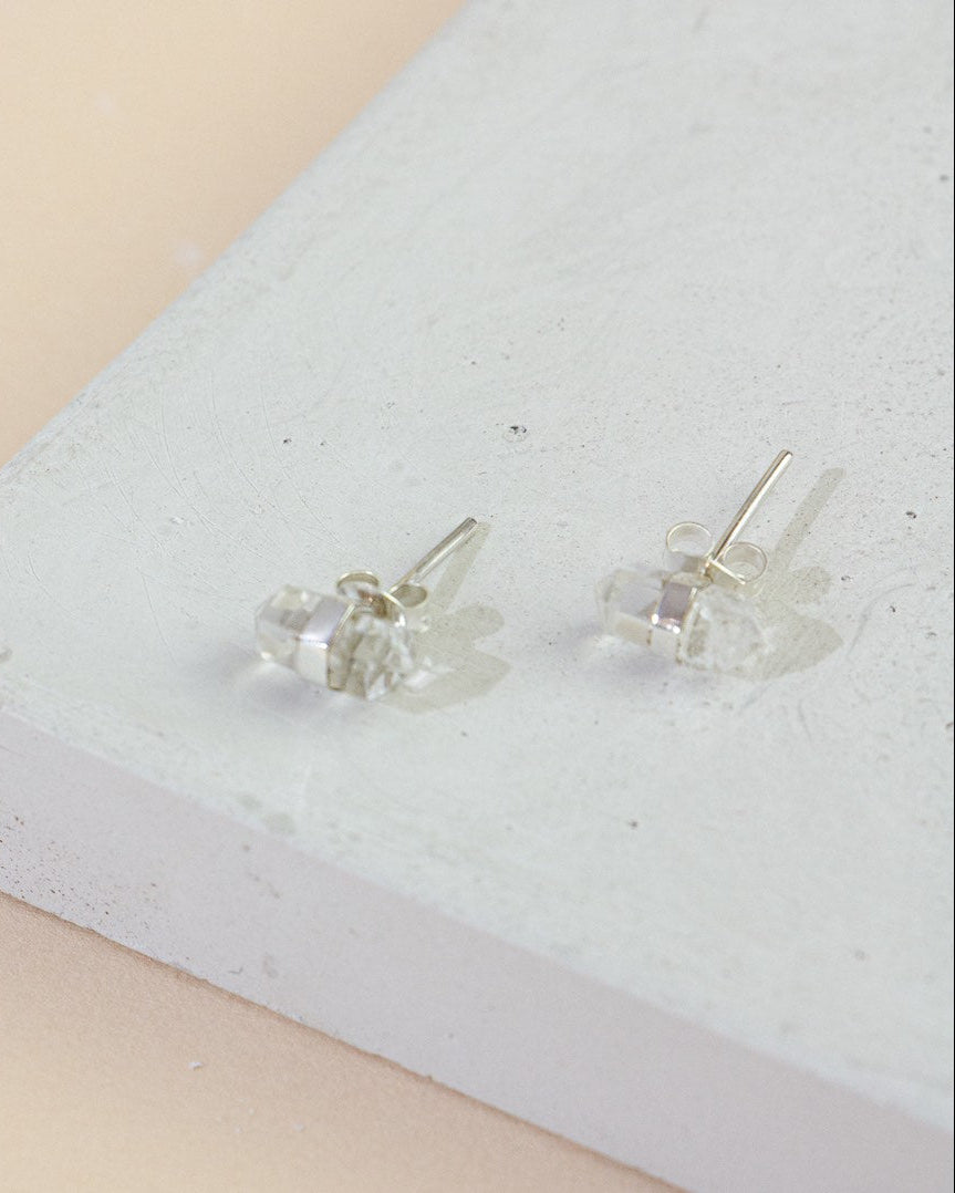 A pair of silver stud earrings featuring a double-terminated clear quartz crystal, displayed on a white surface.